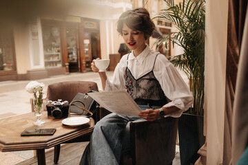 Wall Mural - Beautiful woman in black and white blouse and jeans holds cup of coffee in restaurant. Stylish lady with brunette hairstyle reading menu in cafe..