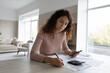 © fizkes - Young Latino woman sit at desk at home manage household expenses expenditure make payment online on smartphone. Hispanic female calculate take care of household budget finances paying on cellphone.