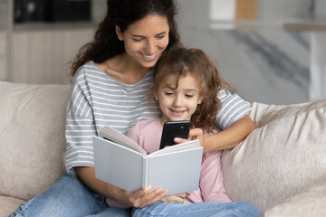 Wall Mural - Happy Hispanic young mother and small daughter relax together read book talk on video call on cellphone. Smiling Latin mom and teen girl child rest at home enjoy story use modern smartphone.