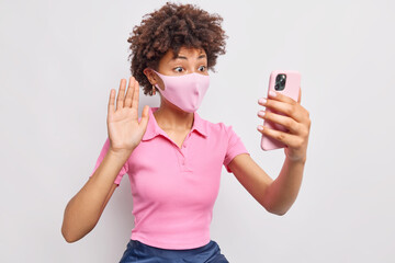 Wall Mural - Afro American woman being on self isolation during coronavirus pandemic wears protective face mask makes video call waves palm in hello gesture keeps smartphone in front poses against white wall