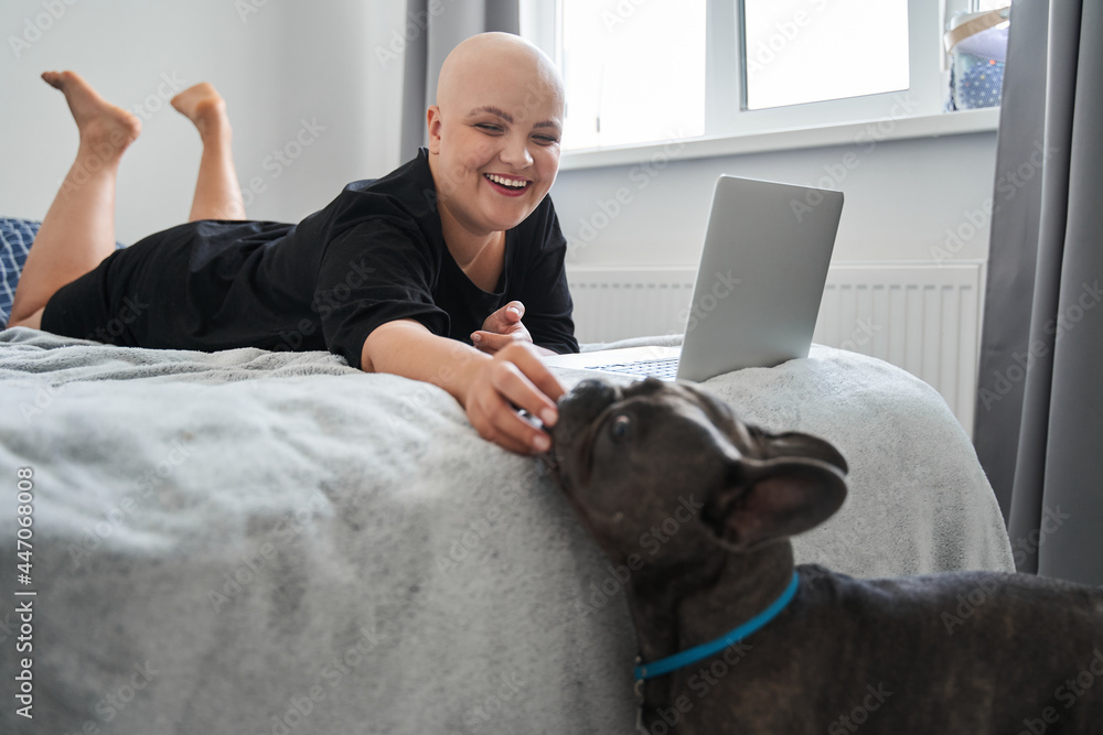 Bald woman feeding her dog while laying at the bed and working - obrazy, fototapety, plakaty 
