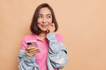 Studio shot of dreamy Asian teenage girl keeps hand on face smiles pleasantly browses internet via smartphone enjoys mobile messaging leisure for chatting poses against brown wall blank space