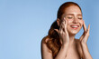 Laughing redhead girl, young woman with flawless skin and freckles, close her eyes from satisfaction, smiling while rubbing, massaging skin with cream, moisturizer or c-serum, blue background