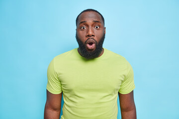 Wall Mural - Portrait of dark skinned man looks embarrassed at camera has shocked expression keeps mouth opened wears casual green t shirt isolated over blue background gasps from fantastic news. Omg no way