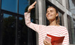 © Liubov Levytska - Happy girl student meeting friend near university campus, standing near building with study books, saying hello. Woman smiling and waving hand to say hi, going on lessons