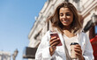 © Liubov Levytska - Beautiful smiling woman walking on street with coffee takeaway, chatting on mobile phone. Girl talking on smartphone on her way to meeting spot, sending message