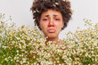 Close up shot of frustrated curly haired Afro American woman surrounded by camomile flowers has red swollen eye srunny nose suffers from seasonal pollen allergy needs consultancy of immunologist