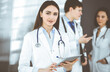© rogerphoto - Smiling intelligent woman-doctor is holding a tablet computer in her hands, while she is standing together with her colleagues in a clinic. Physicians at work. Perfect medical service in a hospital