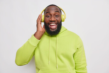 Wall Mural - Handsome bearded hipster man with dark skin enjoys favoite playlist likes listening music keeps hand on wireless headphones smiles broadly dressed in green hoodie isolated over white background