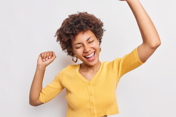 Canvas Print - Positive millennial woman has fun danes with triumph keeps arms raised up moves with rhythm of music wears casual yellow jumper isolated over white background enjoys party celebrates something