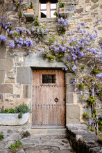 Wooden Door In Old Medieval Stone House, Purple Wisteria In Blossom On The House Over The Door, Rupit, Spain. Magic House From Wonderland