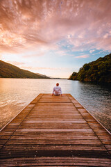 Mergozzo, Verbania / Italy - June 2021: Young man sitting on the wooden jetty of Lake Mergozzo at dawn with colorful clouds in the background