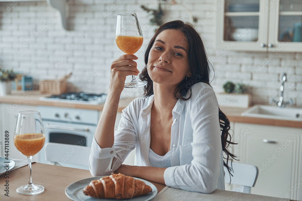 Beautiful young woman holding glass with juice smiling while spending time in the domestic kitchen - obrazy, fototapety, plakaty 