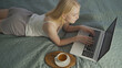 © filin174 - A young woman lies on the bed and communicates on a video call via a laptop. Virtual communication. Comfortable workplace