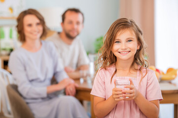 health and beauty concept - smiling little girl holding a glass of water. Happy parents in background.