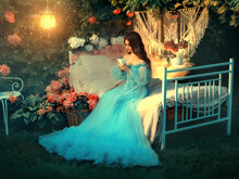 Fairy Tale Princess Sits On White Bed. Fantasy Woman Holding Cup In Hands Drinking Tea. Long Wave Hair. Blue Vintage Luxury Royal Dress. Backdrop Blooming Peony Flowers, Old Lamp. Fantasy Woman Queen.