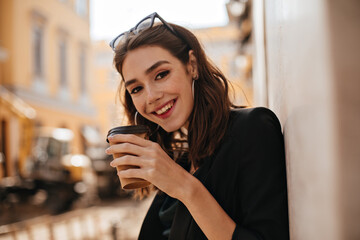 Wall Mural - Cheerful young brunette with wavy hairstyle, trendy makeup in black jacket, smiling, having morning coffee and posing looking into camera at city cafe terrace