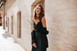© Look! - Pretty young girl with brunette wavy hair, bright makeup, off-shoulder dress and black jacket, holding glasses in hands, walking at daylight city and smiling. Beige building background