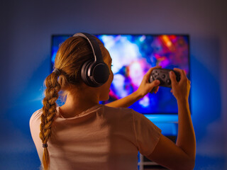 Wall Mural - Woman shooter is playing a video game. Entertainment. Hobby. Leisure. A woman holds a joystick in her hands. She has headphones on her head. Blue neon light pours from the computer monitor.