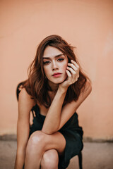 Wall Mural - Well-built young lady with brown wavy hairstyle and trendy makeup in dark slip dress, posing on chair outdoors and looking into camera against peach wall background