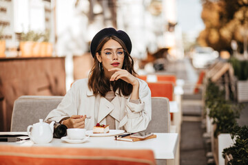 Wall Mural - Attractive young girl in beret, classic beige trench coat, glasses sitting at table with cheesecake and tea. City cafe terrace in sunny autumn