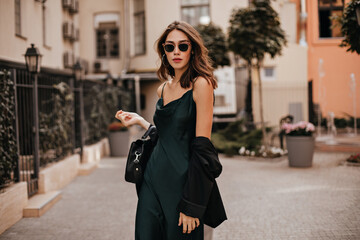fashionable pale brunette in long green dress, black jacket and sunglasses, standing on street durin