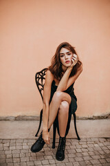 Wall Mural - Full-length portrait of well-built young lady with brunette wavy hair, evening makeup, trending slip dress, black shoes, sitting on chair and crossing legs outdoors
