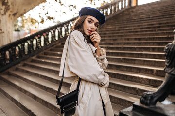 Wall Mural - Lovely young Parisienne with brunette hair in stylish beret, beige trench coat and black bag, standing on old stairs and sensitively posing outdoors. Warm autumn