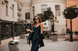 © Look! - Joyful young lady with brunette wavy hair, stylish sunglasses, long silk dress and black bag walking on street, smiling and posing against city architecture background