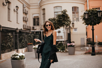 Wall Mural - Joyful young lady with brunette wavy hair, stylish sunglasses, long silk dress and black bag walking on street, smiling and posing against city architecture background