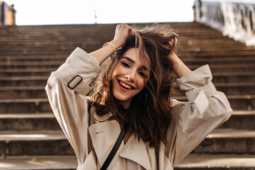 Wall Mural - Cheerful young girl with dark wavy hairstyle and red lips, wearing beige trench, smiling, messing up hair and looking into camera against background of old city stairs