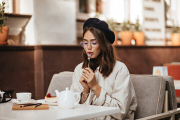 Wall Mural - Charming young dark-haired lady in beret, glasses and beige trench coat having rest at sunny city cafe terrace, eating cheesecake with tea, and looking down