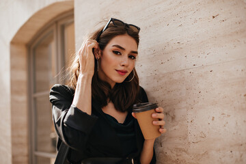 Wall Mural - Pretty young brunette with trendy makeup, glasses on head, dark dress and jacket standing near beige wall in daylight city and holding cup of coffee