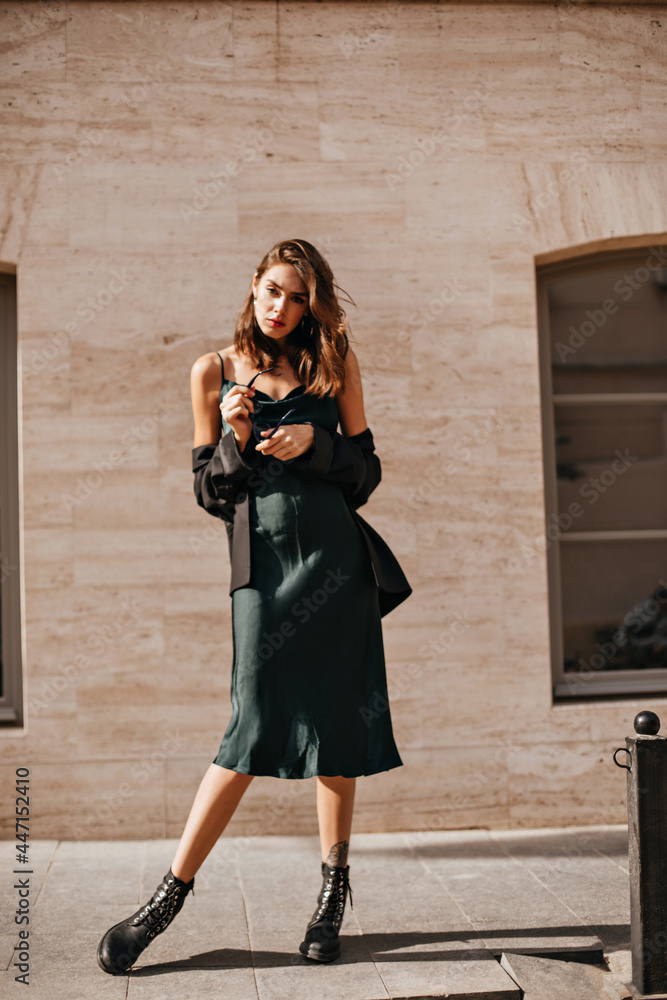 Full-length photo of well-built young girl with dark hair and red lips in  green midi dress, black jacket and shoes, standing and posing against light  building background Stock Photo | Adobe Stock