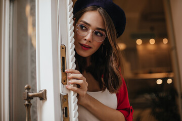 Wall Mural - Cute young girl with dark hair, red lips and shirt, wearing beret on head, glasses peeping out white door against golden background