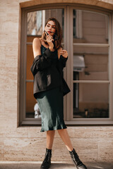Wall Mural - Elegant young woman with brunette hair, red lips, stylish dress, black jacket and boots, posing outdoors, holding sunglasses and looking away against beige wall and big window background