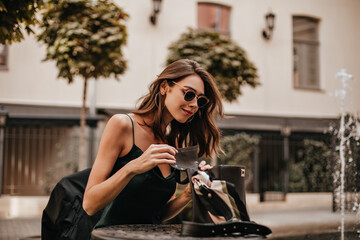 Wall Mural - Charming young lady with brunette wavy hair, trendy sunglasses, green silk dress sitting outdoors on city cafe terrace, opening black bag and smiling