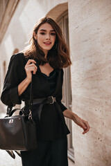 Wall Mural - Charming young brunette with wavy hairstyle, nude makeup, dark dress, black jacket and bag, stylish belt on waist, posing against beige wall background