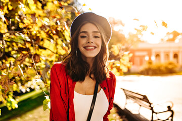 Wall Mural - Gorgeous young girl with medium dark hair, white top, red shirt and beret, smiling and posing looking straight into camera. Warm autumn city background