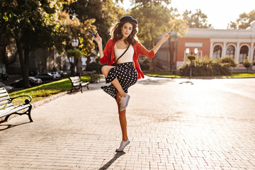 Wall Mural - Lifestyle portrait of active young brunette in French style outfit posing, lifting knee and hands outdoors. City square during warm sunny day