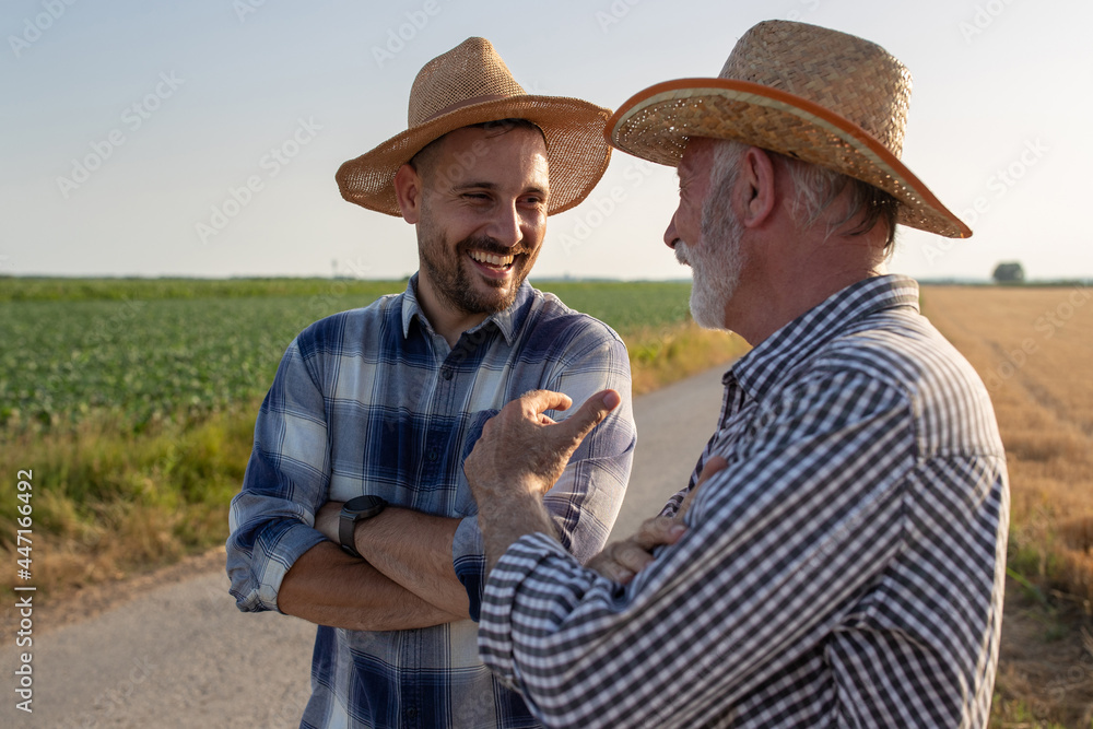 Two farmers standing on road next to field talking laughing. - obrazy, fototapety, plakaty 