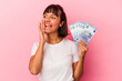 © Asier - Young mixed race woman holding bills isolated on pink background shouting and holding palm near opened mouth.