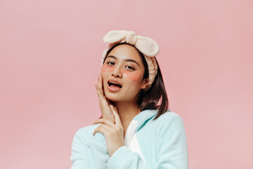 Wall Mural - Pretty young woman in soft blue pajamas and beige headband touches her face, looks into camera and poses with cosmetic eyepatches.