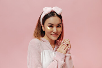 Wall Mural - Charming brown-eyed woman in headband and kigurumi smiles and poses with eyepatches on pink background.