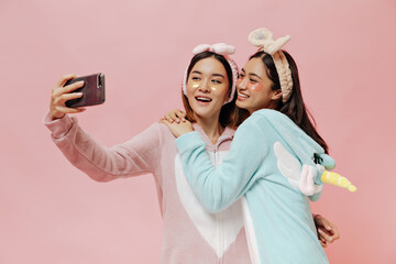 Wall Mural - Young attractive Asian women in pajamas take selfie on pink background. Happy brunette girls in headbands hug on isolated.