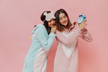 Wall Mural - Cute short-haired girls in soft pajamas pose on pink background. Young girls in kigurumi holds blue alarm clock on isolated.