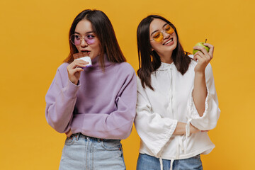 Wall Mural - Pretty girl in purple sweater bites milk chocolate bar. Young woman in white hoodie looks at green apple and smiles. Portrait of charming Asian women pose on orange background.