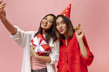 Wall Mural - Tanned brunette Asian women take selfie and celebrate birthday on pink background. Cute girl in white shirt poses with red gift box. Charming lady in party hat holds party horn.