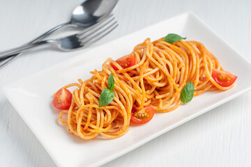Wall Mural - Plating of delicious italian spaghetti pasta portion cooked in red sauce decorated by chef with green basil leaves and cherry tomatoes served on rectangular plate on white wooden background with fork