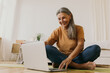 © shurkin_son - Indoor image of beautiful energetic female on retirement sitting barefoot on floor using laptop turning on yoga video tutorial. Elderly European woman surfing internet on portable computer